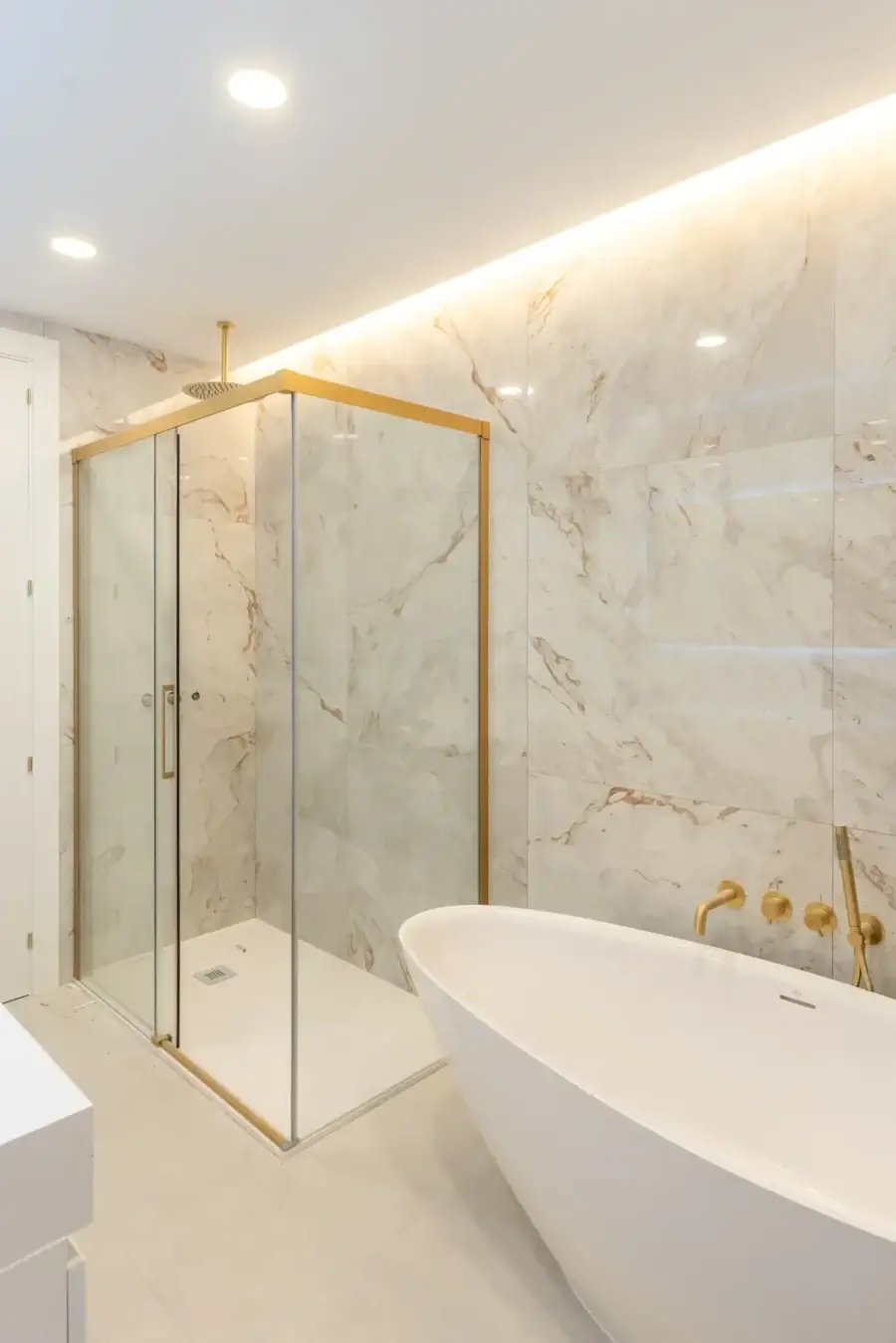 Baño reformado con revestimiento de mármol claro, mampara de vidrio y grifería dorada
